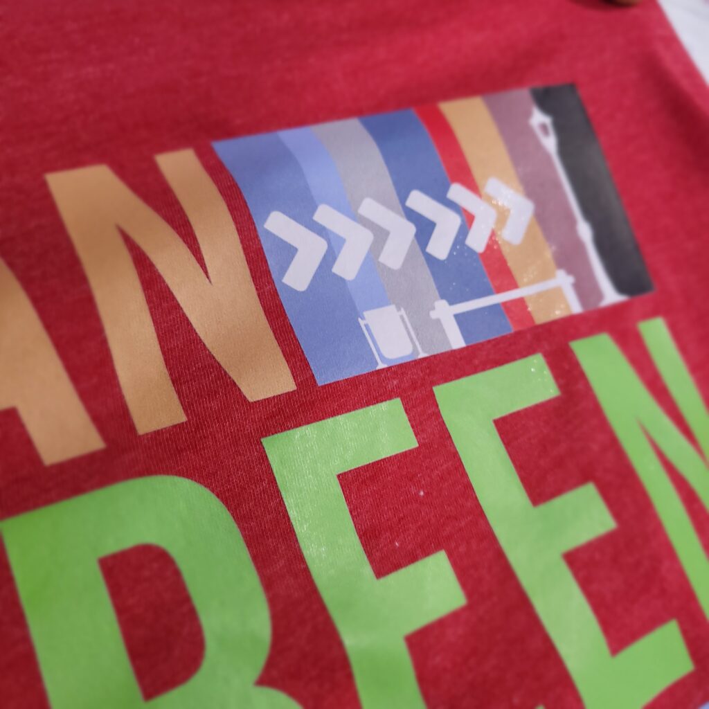 Close-up of a colorful graphic design on a red surface, featuring abstract shapes including arrows and silhouettes. Large, partially visible green and orange text is displayed beneath the design.